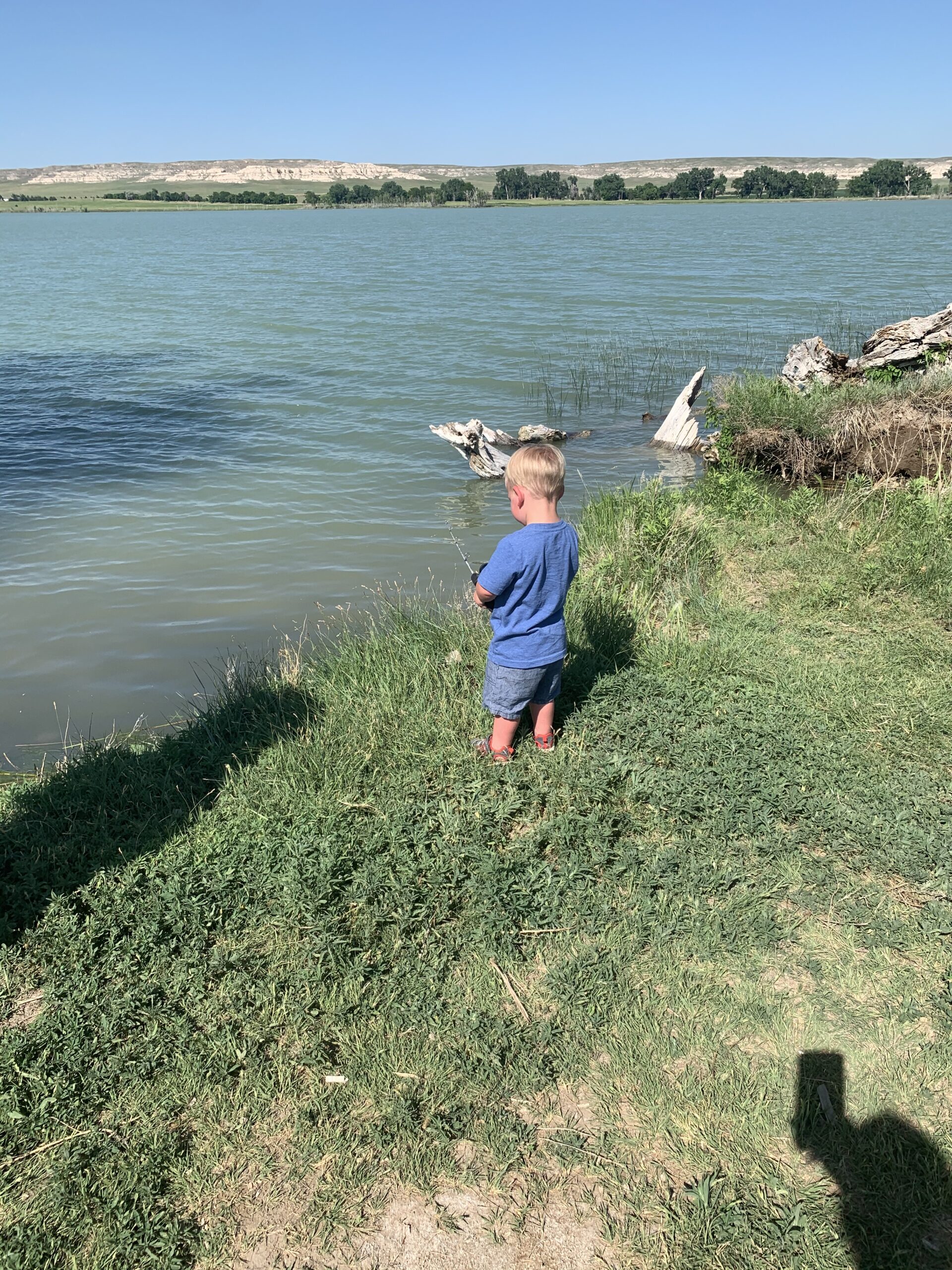 A little boy is standing on the shore of a lake fishing with his fish rod in the lake.