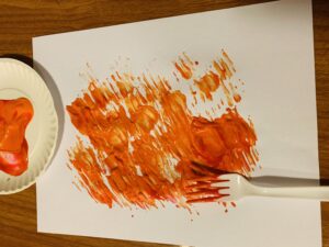 A white paper is the main part of the image. On the white paper there is red, orange, and yellow paint that has been painted with a fork. The fork is laying on the right side of the paper. On the left side of the paper is the paper plate the paint was mixed on before painting.