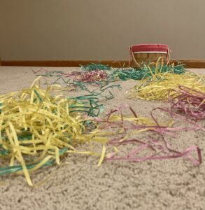 There is Easter grass all over the floor of a room. Behind the Easter grass sits an Easter basket.