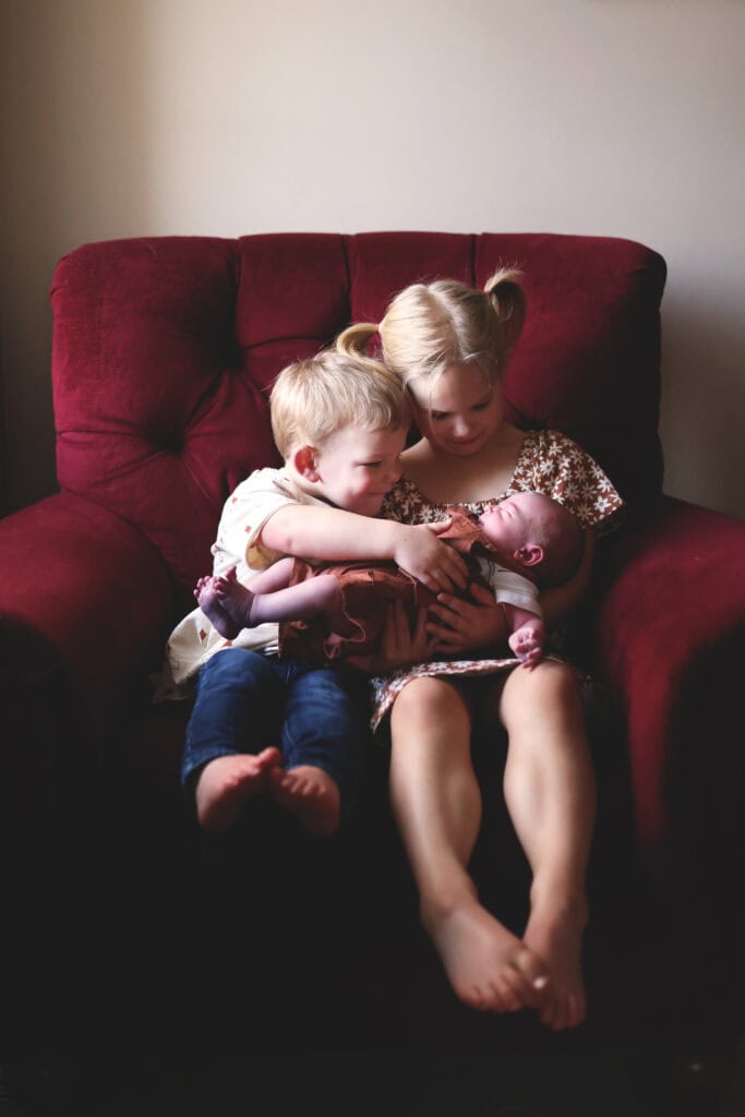 Two young siblings sitting on a red armchair, lovingly holding their newborn baby sister—capturing the sweet sibling bond and real-life moments that unfold while homeschooling with a newborn.