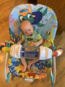 Newborn baby sitting in a colorful ocean-themed baby bouncer with toys, smiling and engaged. A playful and practical setup for parents who are homeschooling with a newborn.