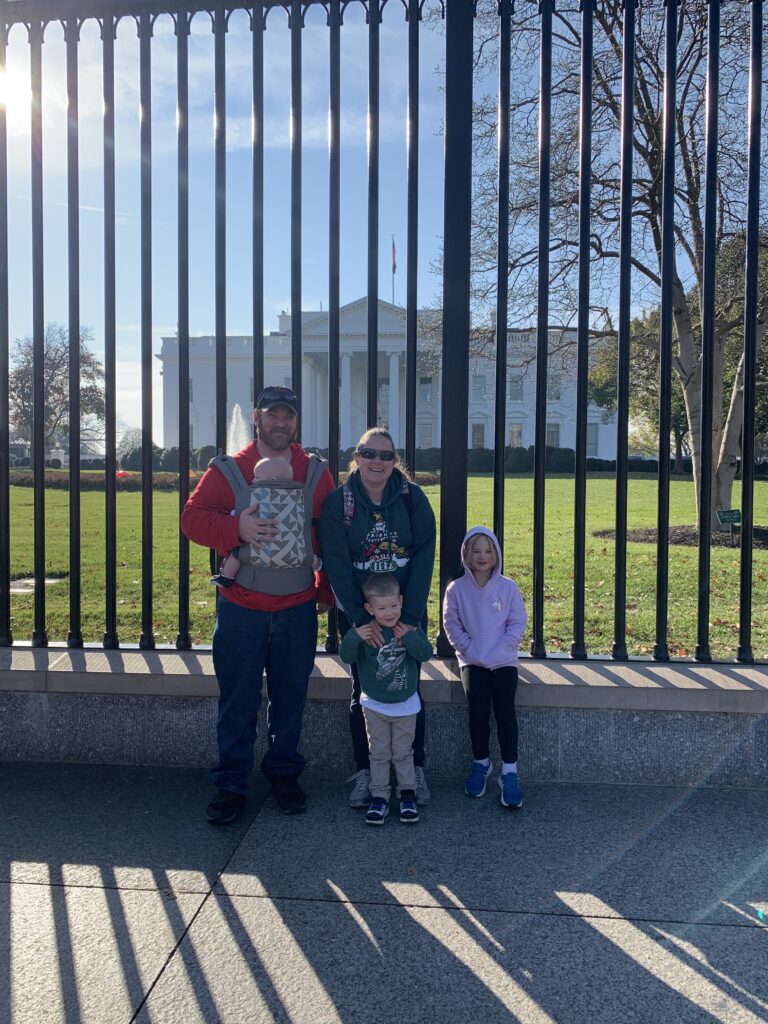Family of five standing in front of the White House gates on a sunny day, with the father wearing a baby in a patterned wrap carrier—showcasing how babywearing supports flexibility while homeschooling with a newborn during family outings and educational travel.