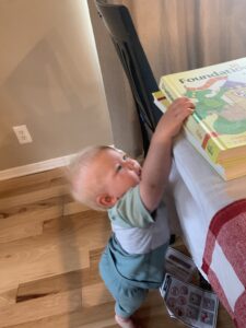 Curious baby reaching up to grab homeschool curriculum books from a dining table, highlighting the everyday challenges and multitasking involved in homeschooling with a newborn or toddler in the home.