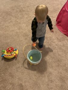 A toddler boy is placing an Easter egg into a basket during an egg hunt.
