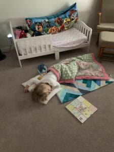 Toddler girl asleep on the carpet with a quilt and pillow during homeschool rest time, with a toddler bed and children’s book nearby—capturing a peaceful moment while homeschooling with a newborn and young children.