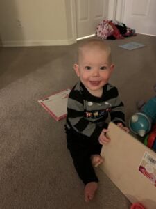 Smiling baby sitting on a carpeted floor holding a wooden puzzle board, surrounded by toys and books—a joyful moment during a typical day of homeschooling with a newborn and toddler at home.