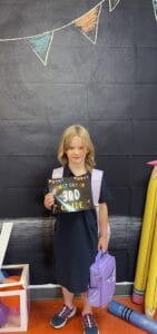 Smiling child holding a 'First Day of 3rd Grade' sign with a backpack, standing in a decorated classroom area—showcasing how hybrid or outside schooling can help outsource education while homeschooling with a newborn at home.