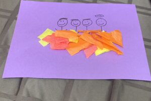 Tissue paper art project A purple sheet of construction paper is the main image. On the paper there are three boys and one angel drawn. On top of the drawings, there is red, yellow and orange tissue paper glued.