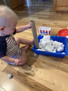 Young toddler boy playing with a cotton ball sensory bin. He is holding a paper towel roll inside a blue sensory bin full of cotton balls with a bowl and a bucket behind it. This idea was found in the Toddler Play & Grown Activity Guide.
