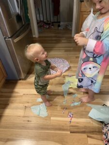 A young toddler boy is standing and staring at his older sister who is tearing tissue paper. They are surrounded with tissue paper on the floor. This activity is found in the Toddler Grow & Play Activity Guide.