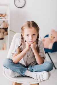 Young girl with braids sitting cross-legged on a chair, looking upset—highlighting the importance of emotional awareness from the social emotional skills checklist for a homeschool rising kindergartener.