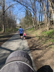 A family walks along a wooded path on a sunny day, with a preschool-aged child holding hands with an adult and a stroller in the foreground. This outdoor learning preschool moment encourages exploration, movement, and nature observation.