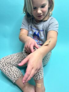 Little girl is sitting on a blue background putting bandaids on her body for to show one of the Bible activities for toddlers or preschoolers.