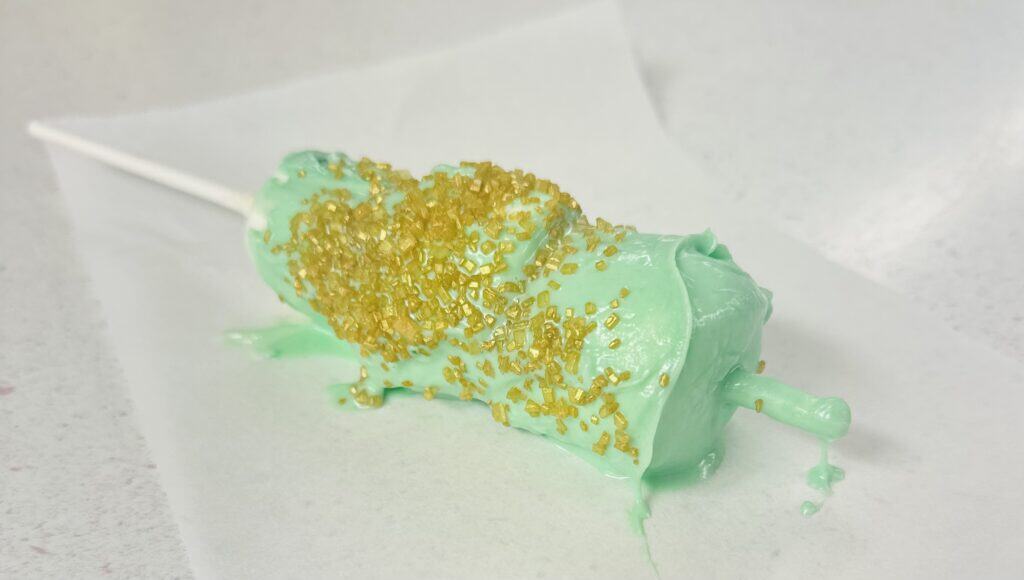 A set of three marshmallows stuck on a stick covered in green chocolate with gold sprinkles is laying on a piece of parchment paper on a counter.