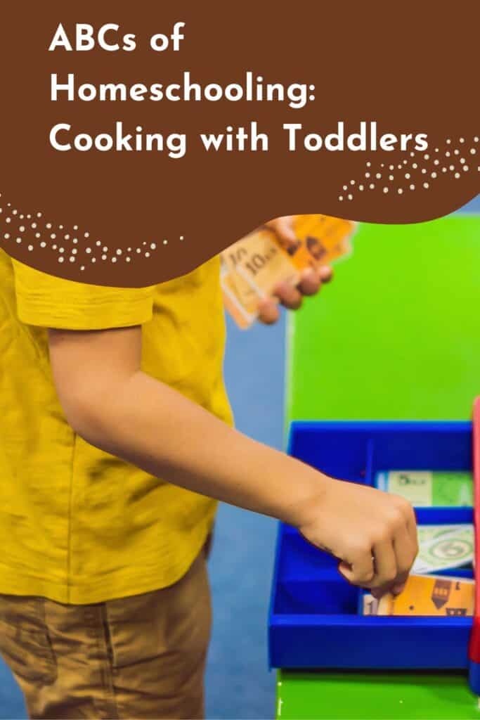 Pinterest image for "ABCs of Homeschooling: Cooking with Toddlers" featuring a child in a yellow shirt using play money and a toy cash register. This image highlights pretend play, early math skills, and hands-on toddler learning through dramatic play and role-based activities. Ideal for homeschooling and educational play ideas.