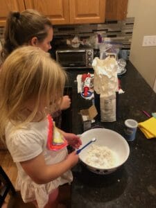 A young child holds a measuring spoon over a large mixing bowl filled with flour, while an adult stands nearby offering guidance. Baking powder, flour, and other ingredients are spread out on the kitchen counter. This candid moment highlights the learning and connection that happens during cooking with toddlers.
