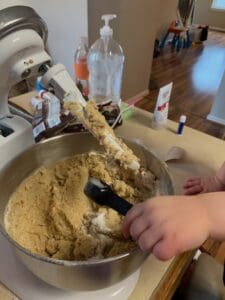 A toddler's hand adds a spoonful of flour to a large mixing bowl of cookie dough in a stand mixer. The countertop is scattered with baking supplies, and the scene captures the mess and joy of cooking with toddlers in a real-life kitchen setting.