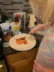 A young child spreads sauce onto a tortilla using a spoon, standing at a kitchen counter surrounded by ingredients like yogurt, canned black beans, and tortillas. This hands-on moment captures the real-life experience of cooking with toddlers and encouraging independence in the kitchen.