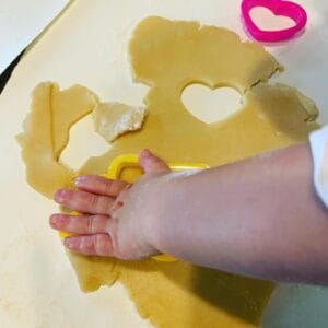 A toddler presses a yellow heart-shaped cookie cutter into rolled-out sugar cookie dough on a floured surface. Another pink cookie cutter and a heart-shaped cutout are visible nearby. This sweet moment captures the hands-on joy and sensory learning that comes with cooking with toddlers.