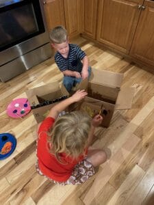 wo young children sitting on a hardwood kitchen floor painting and decorating a cardboard box, using colorful paint palettes and brushes. This image captures pretend play, creativity, and hands-on learning for toddlers and preschoolers, promoting imaginative play in a homeschool or home environment.