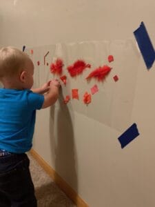 Toddler engaging in a red-themed sticky wall activity by placing feathers and paper onto contact paper—an interactive way to build fine motor skills through color sorting and sensory play.