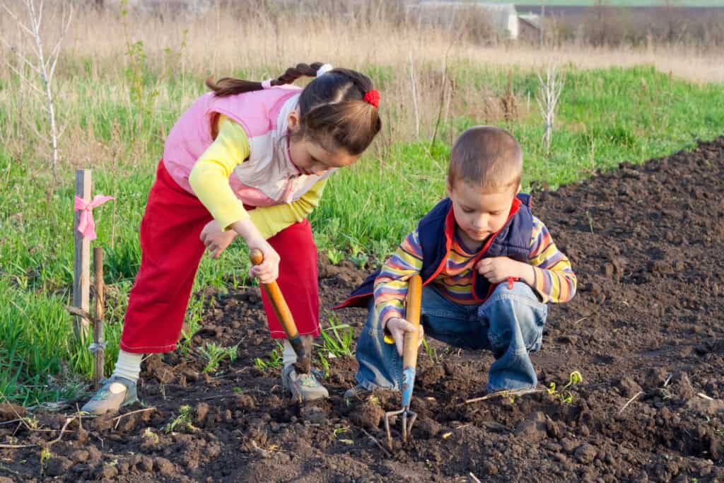 Two young children working in a garden with small tools, learning responsibility and kindness through gardening as a way of teaching kids to serve others