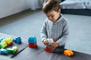 A toddler wearing a light gray cable-knit sweater is standing indoors, focused on stacking large colorful plastic building blocks on a gray ottoman. More blocks are scattered nearby. The room has a cozy, carpeted floor with a bed visible in the background.