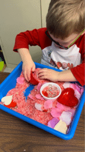 Preschool child wearing glasses and a red shirt scooping pink rice from a Valentine-themed sensory bin using a red silicone baking cup. The bin contains dyed rice, artificial flower petals, heart-shaped containers, and clear gems—showcasing creative, hands-on sensory bin products for themed sensory play.