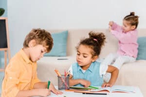 Two young children working with colored pencils at a table, engaging in creative learning activities that support kindergarten readiness through play, while another child plays in the background.