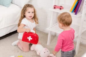 Young girl playing doctor with a toy medical kit and stuffed animal, while another child watches—modeling imaginative play that supports kindergarten readiness through play.