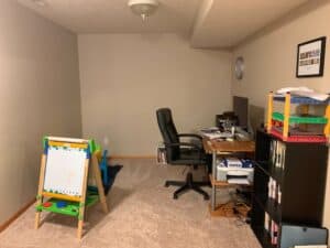 A basement homeschool room set up for a homeschooling toddler preschooler. On the left, a child’s easel with a whiteboard and colorful magnets sits on the carpet. Behind it is a small blue table and chairs for kids. On the right, a computer desk holds papers, a monitor, and supplies, with a black bookshelf beside it filled with books and a colorful toy shelf on top. The space is organized for both adult work and early childhood learning activities.