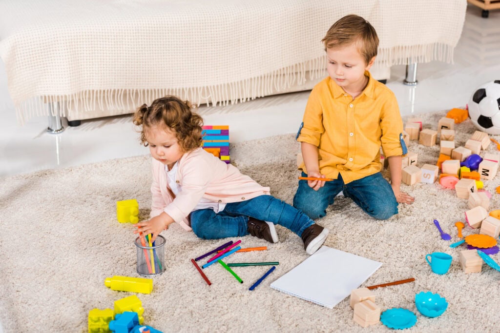 Two young children sitting on a rug surrounded by colorful toys, blocks, and crayons, engaging in creative play to teach letters to toddlers and preschoolers through hands-on learning.