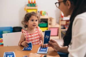 Toddler in a pink checkered dress holding a letter M flashcard while learning with a teacher who is showing the letter P, engaging in a hands-on activity to teach letters to toddlers and preschoolers.