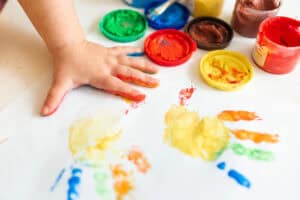 A young child’s hand pressing into colorful finger paint to make handprint art on white paper, surrounded by open paint jars—capturing a classic example of easy toddler activities at home that build creativity and sensory skills through messy play.