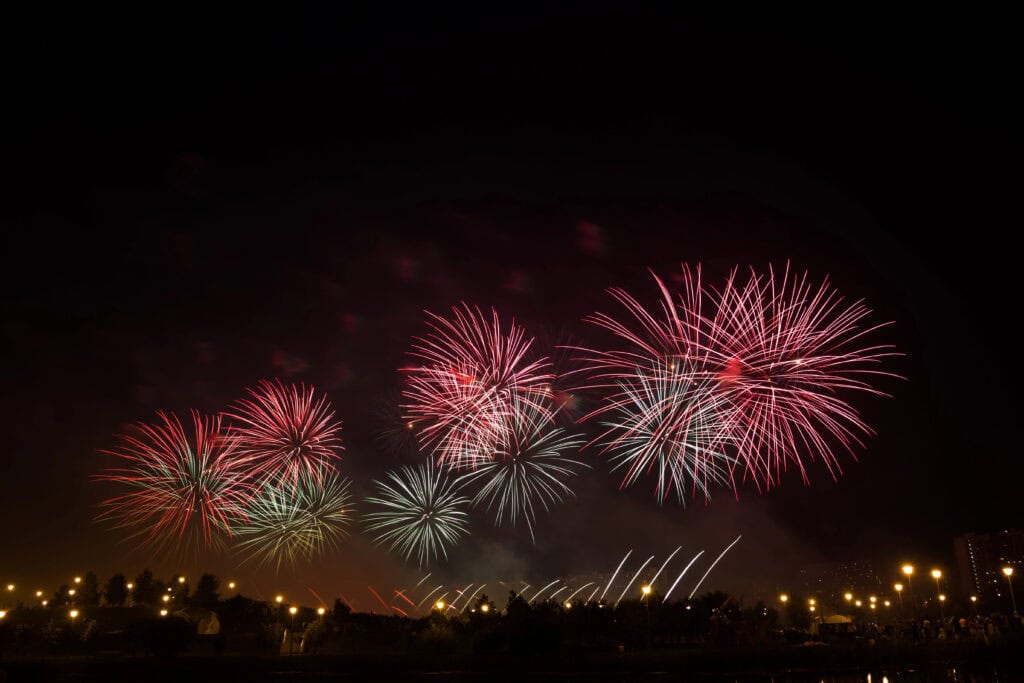 Bright red, white, and colorful fireworks exploding across a dark night sky above a park with glowing streetlights and silhouettes of trees and people watching the celebration below.