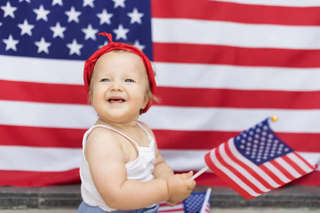 Smiling baby wearing a red headband and holding a small American flag while sitting in front of a large United States flag backdrop