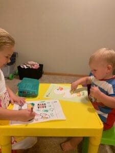 Two young children sitting together at a child-sized desk, coloring homeschool worksheets with crayons in a playroom.