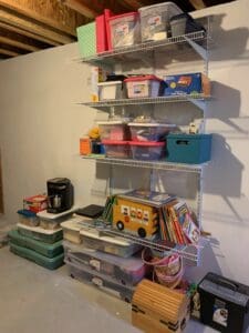 Homeschool organization area with wire shelving, clear storage bins, books, learning toys, games, and school supplies stored neatly in a basement.